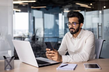Ciddi Müslüman genç adam ofiste çalışıyor, masada oturuyor ve telefon kullanıyor, mesajlaşıyor, faturaları kontrol ediyor, telefon görüşmeleri yapıyor..