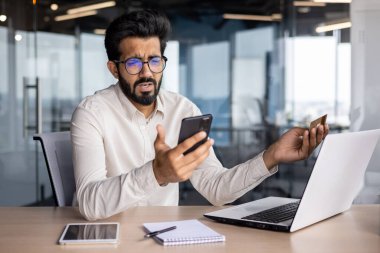 Hesapta sorun var. Ofiste çalışan üzgün ve endişeli genç Hintli işadamı elinde kredi kartıyla telefon ekranında hayal kırıklığına uğramış görünüyor..