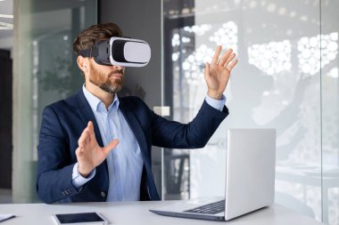 Ofiste çalışan genç bir adam, bilgisayarlı bir masada oturuyor, sanal bir maske takıyor ve video ile uzaktan konuşuyor, elleriyle hareket ediyor..