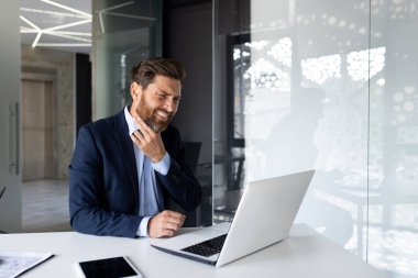 Genç bir erkek işadamı ofiste bir masada oturup boğazını tutuyor. Acı ve buruşukluk hisseder..