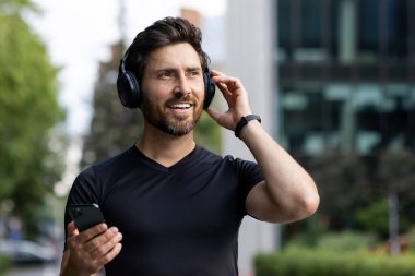 Şehir caddesinde duran, kulaklık takan, elinde telefon tutan ve gülümseyen genç bir atletin yakın plan fotoğrafı..