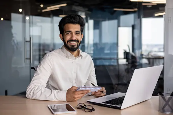 Portrait of a young Indian successful man smiling and looking at the camera, sitting in the office at a desk with a laptop, holding a notebook.