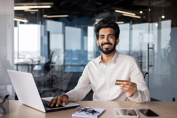Portrait of a smiling young Indian man financier holding a credit card, working on a laptop and conducting banking transactions.