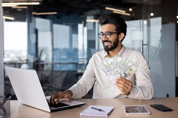 Indian smiling young man businessman working in the office on a laptop, sitting at the table and holding cash bills in his hand.