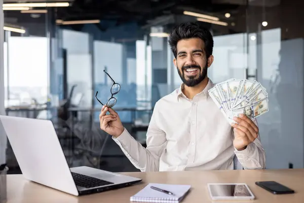 Portrait of a young happy Indian male businessman sitting in the office at the table with a laptop and rejoicing in financial success, holding a fan of money banknotes and glasses in his hand.