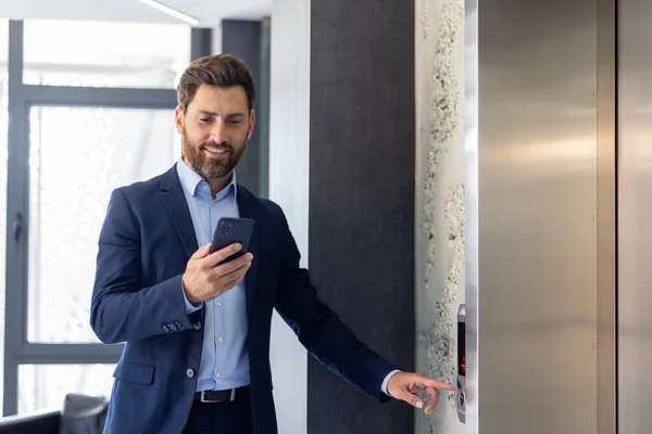A smiling young male businessman is standing in an office space, calling an elevator and using a mobile phone.