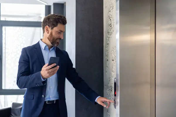 A smiling young man businessman is standing in an office space near the elevator and presses a call button, holding a phone in his hands.