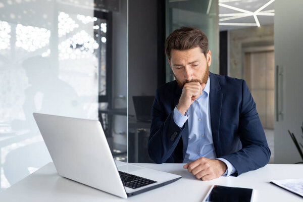 A young male businessman sits in the office at the desk, is sick and coughs, covers his mouth with his hand.