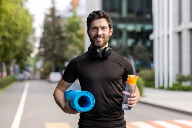 Bir şehir caddesinde elinde bir paspas ve bir şişe su tutan gülümseyen genç bir erkek atletin yakın plan portresi. Durmadan kameraya bakıyor..