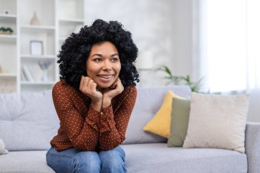 Gülümseyen ve mutlu Afro-Amerikan genç kadın evinde kanepede oturuyor, ellerini dizlerinin üstüne koyuyor, hayaller kurarak ve düşünceli bir şekilde gözlerini kaçırıyor..