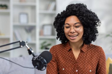 Evinde mikrofonla podcast yapan neşeli genç bir kadın, sıcak bir iç mekan ortamında iyimserliğini ve kendine güvenini ifade ediyor..