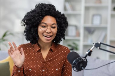 Afro-Amerikalı genç bir kadın evde oturuyor ve mikrofon aracılığıyla konuşuyor, podcast, uzaktan radyo, el sallama ve selamlama kaydediyor. Yakın plan fotoğraf..