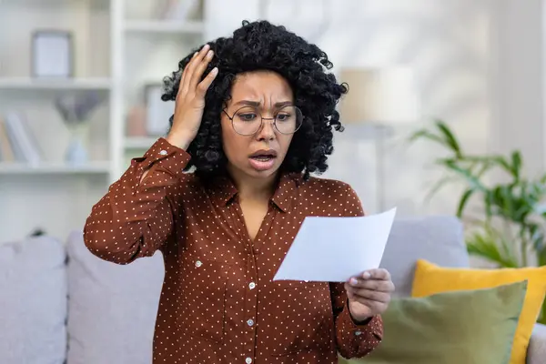 Close-up photo of a shocked young African American woman at home, looking shocked and holding documents, messages, worriedly holding her head.
