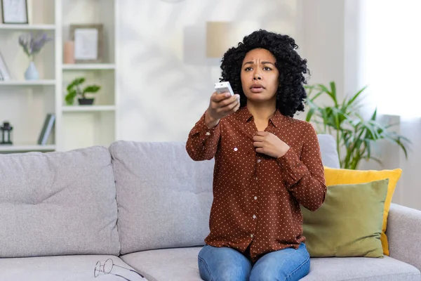 Worried young African American woman sitting on sofa at home and turning on air conditioner by remote control. He feels bad.