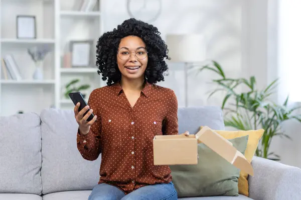 Portrait of happy African American woman sitting on sofa at home, holding open parcel box and mobile phone, looking and smiling at camera.