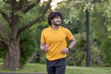 Gülümseyen genç Hintli adam parkta koşuyor, sabah koşuyor ve egzersiz yapıyor..