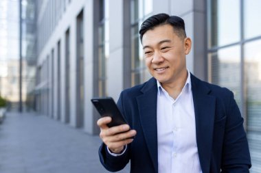 Asyalı gülümseyen genç adam bir ofis binasının yanında yürüyor ve cep telefonu kullanıyor..