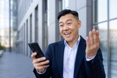 Ofis merkezinin önünde duran Asyalı genç bir iş adamının yakın plan fotoğrafı. Telefon ekranına bakıyor ve duygusal olarak mutlu..