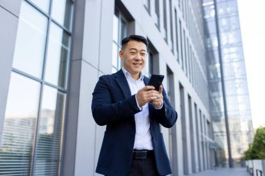 Asyalı genç işadamı ofis binasının önünde duruyor ve cep telefonu kullanıyor. Görüşmeyi bekliyor, arıyor ve mesaj yazıyor..