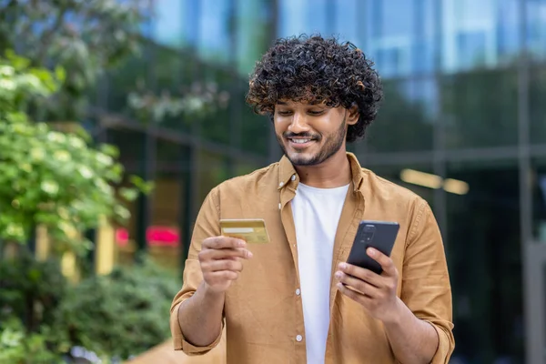 Indian smiling young man standing on city street, holding phone, looking and using credit card.