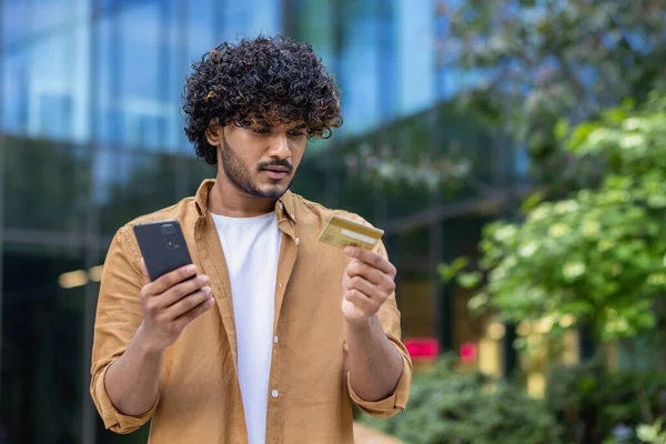 Closeup photo of a worried young Muslim man holding a phone and looking confusedly at a credit card.