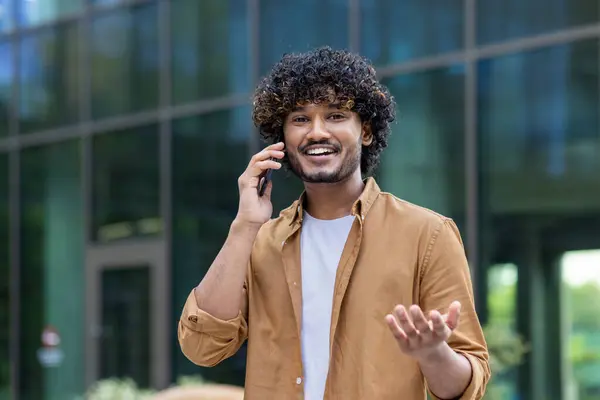 Close-up portrait of a young smiling Muslim man standing on the street and talking on the phone, looking and gesturing at the camera.