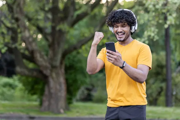 A young Indian sportsman checks his phone and celebrates while listening to music in the park after a run.