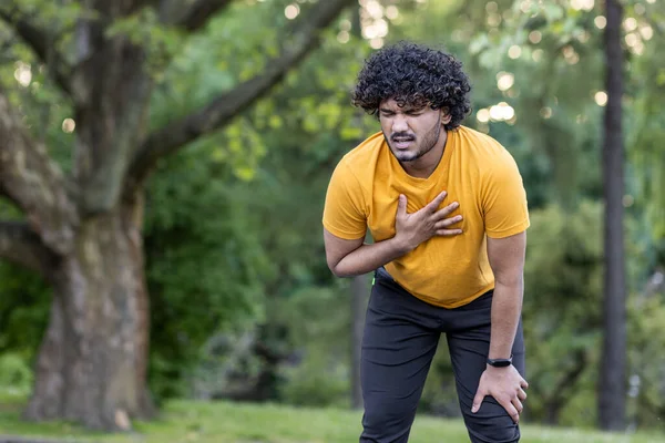 Tired young Indian male athlete standing bent over in park and holding hand to chest, feeling severe pain and shortness of breath after jogging.