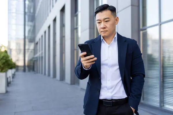 Serious young Asian man standing outside an office building in a suit, holding his hand in his pocket and using a mobile phone.