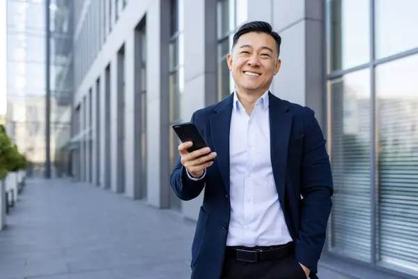 Portrait of a young Asian male businessman standing smiling on the street near an office center, holding a phone and looking at the camera.