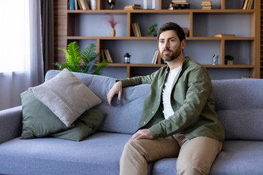 Evdeki kanepede oturan, ciddi ve günlük gömlekle düşünen, oturma odasında dinlenen olgun bir adam..