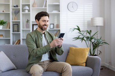 Oturma odasındaki koltukta oturan tatmin olmuş olgun bir adam, telefondaki uygulamayı kullanarak, akıllı telefonu tutan serbest çalışan, mesaj yazan ve okuyan, sosyal medyada gezinen.