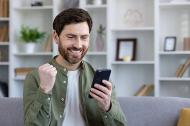 Çevrimiçi galibiyet bildirimi almış ve başarılı online başarı sonuçları telefonda, evdeki koltukta oturuyor, kazanan elini kaldırıyor, zafer işareti yapıyor..