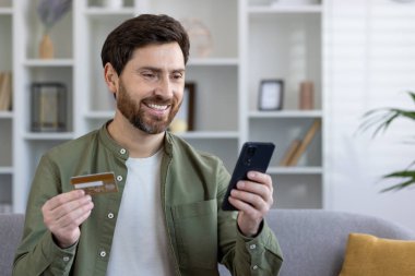 Evdeki koltukta oturan bir adamın internetten online alışveriş için başvuru formu, kredi kartı ve banka kartı kullanması tatmin edici bir şekilde gülümsemesi, hediyeler seçmesi..