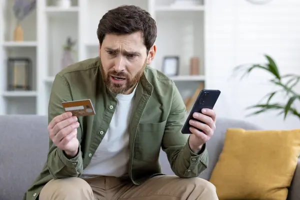 Upset cheated man sitting on couch in living room at home, holding bank credit card and phone, rejected money transfer, negative balance, scam money loss.