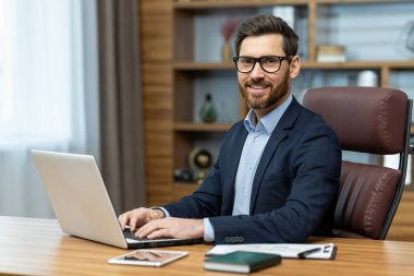 Şık takım elbiseli, gülümseyen çekici bir adam bilgisayarın klavyesinde el ele tutuşuyor ve kendine güvenerek kameraya bakıyor. Başarılı çalışan şirket ve kişisel kariyer başarılarından memnun..