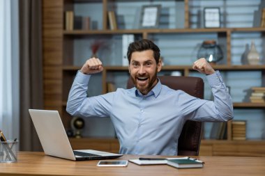 Mavi gömlekli, mutlu sakallı bir erkek bilgisayar başında çalışırken yumruklarını sıkıp el kaldırıyor. Neşeli girişimci sevinç çığlıkları atarken ve başarılı bir günü kutlarken hisseder..