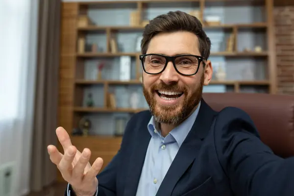 First person view of cheerful adult man in glasses having video call with happy facial expression on luxury interior background. Excited office worker presenting new workplace with toothy smile.