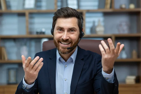 First person view of handsome man in formalwear and headset smiling confidently and gesturing actively during video call. Startup owner making business presentation for new investors attraction.