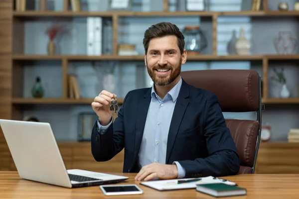 Happy attractive man in shirt and jacket sitting by desktop with laptop and holding keys in hand. Smiling property developer feeling delighted about profitable deal with work partner or client.