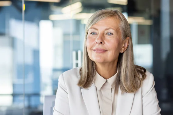 Close-up portrait of a successful and self-confident senior gray-haired business woman sitting in the office at the workplace and looking away with a smile.