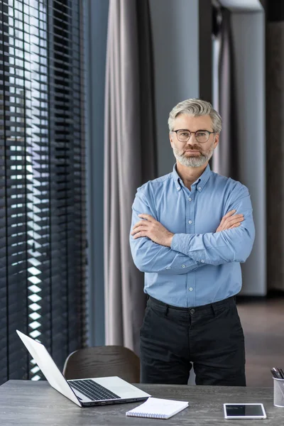 Serious grey haired boss in eyewear and formal clothes standing by desktop with laptop with crossed arms on chest. Strict head of company preparing for speech about deadlines in front of employees.