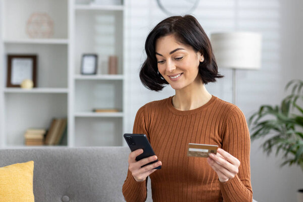 Улыбающаяся молодая женщина сидит дома на диване, держит кредитку и пользуется мобильным телефоном. Крупный план.