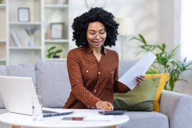 Uzaktan kumandalı iş. Afro-Amerikalı bir kadın evdeki evrak işlerini yapıyor, verileri kontrol ediyor, hesap makinesi kullanıyor..