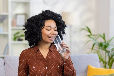 Afro-Amerikalı genç kadın bardaktan su içiyor, evdeki kanepede gülümsüyor, susuzluğunu gideriyor, su dengesini koruyor. Yakın plan fotoğraf..