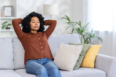 Yorgun ve ciddi bir Afro-Amerikalı kadın. Kanepede oturuyor ve dinleniyor. Elleri başının arkasında ve gözleri kapalı..