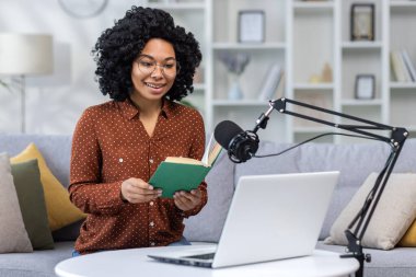 Gülümseyen Afro-Amerikan kadın evdeki koltukta oturuyor mikrofon ve dizüstü bilgisayarla, video bağlantısıyla kitap okuyor..