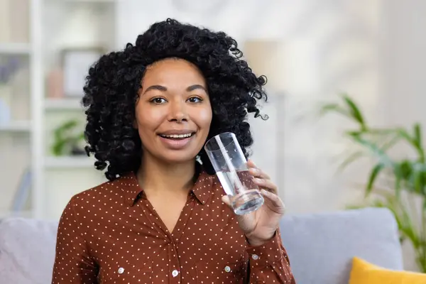 Evdeki kanepede oturan ve elinde bir bardak temiz su tutan Afro-Amerikalı genç bir kadının yakın plan portresi. Kameraya bakarken mutlu oluyorum..
