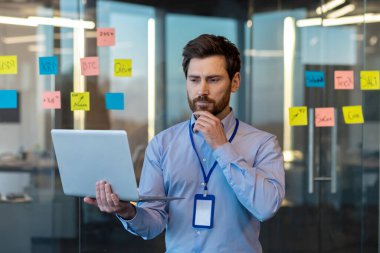 Düşünceli ve konsantre genç adam ofiste çalışıyor, elinde bir dizüstü bilgisayar tutuyor, ciddi bir şekilde ekrana bakıyor, bir probleme ve bir iş sorununa çözüm düşünüyor..