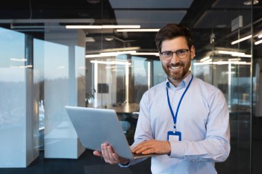 Gülümseyen genç bir ofis çalışanının portresi, uzman bir iş adamı pencerede gömlek içinde duruyor ve rozet takıyor, dizüstü bilgisayarını tutuyor ve özgüvenle kameraya bakıyor..